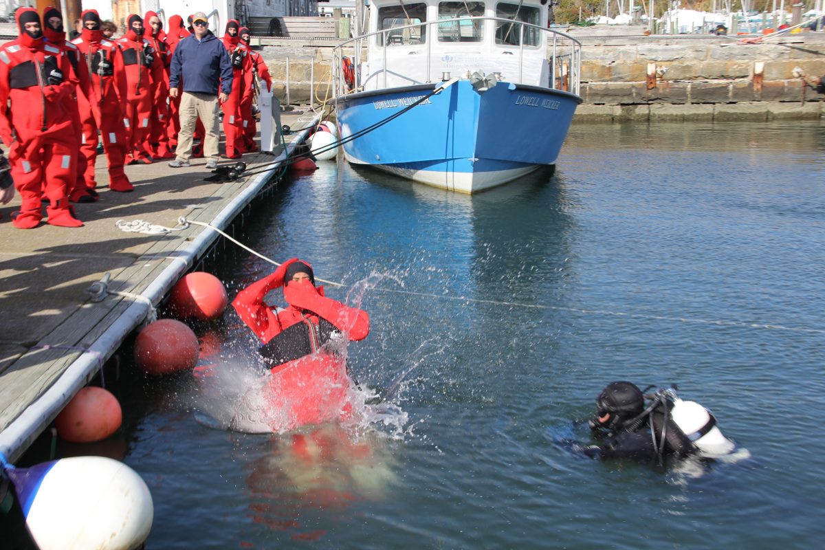 A fisherman enters the water in his immersion suit, with a safety diver on standby nearby.